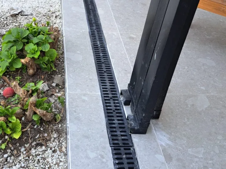 Rejilla lineal negra colocada junto a un piso gris, al pie de una columna oscura y cerca de un cantero.