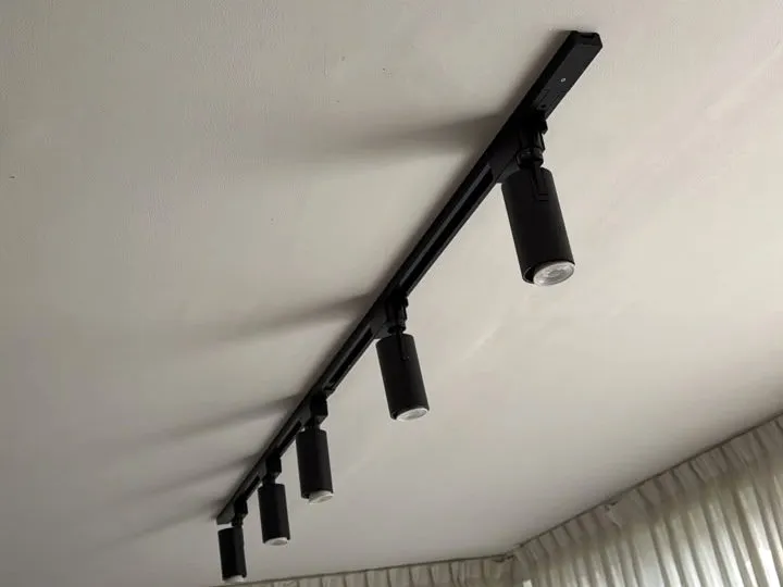 Tramo de riel negro con spots orientables instalado en un techo blanco, junto al borde de unas cortinas.