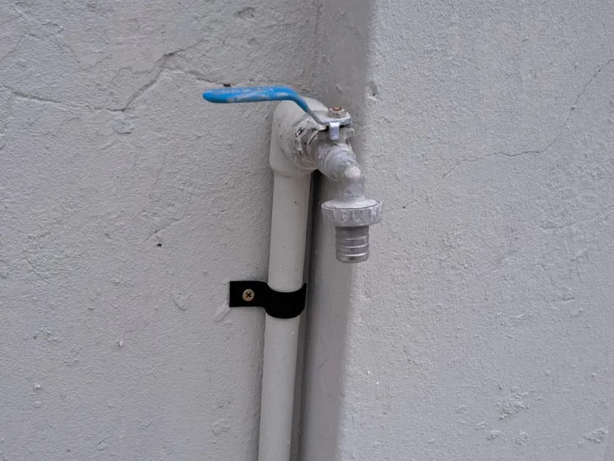 Canilla exterior de palanca azul con cañería blanca a la vista y abrazadera negra sobre una pared gris.