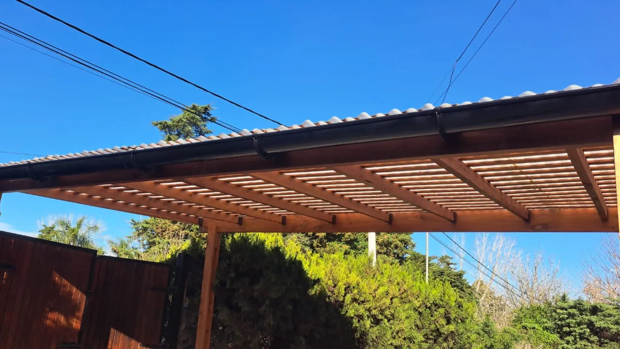Pérgola de madera con vigas y listones, cubierta clara y canaleta oscura, junto a vegetación del patio.
